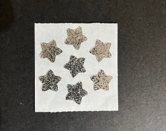 May include: A square piece of white paper with seven glittery star stickers arranged in a cluster. The stars have a silver and black speckled appearance, and the paper has a rough, torn edge.