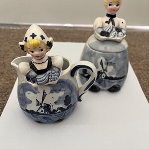 May include: A ceramic creamer and sugar bowl set. The creamer is a small pitcher with a handle, and the sugar bowl has a lid. Both are decorated with blue and white windmill scenes and figurines of people in traditional Dutch attire.