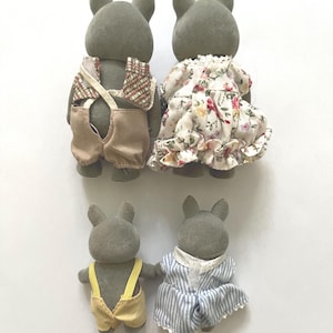 Pu&ograve; includere: Quattro statuette di conigli grigi. Due conigli adulti sono vestiti con un gilet a quadri e una salopette beige, e un vestito floreale. Due coniglietti pi&ugrave; piccoli indossano una salopette gialla e un vestito a righe blu.