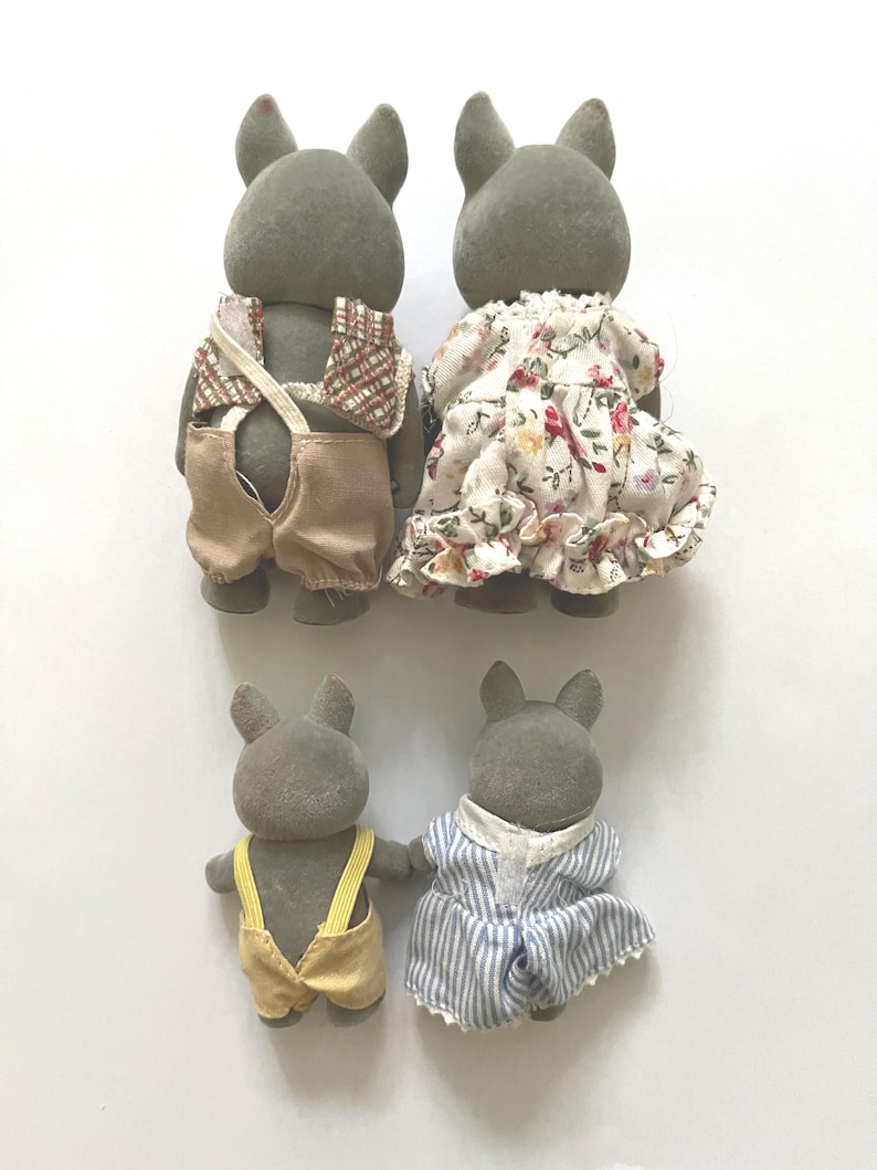 Pu&ograve; includere: Quattro statuette di conigli grigi. Due conigli adulti sono vestiti con un gilet a quadri e una salopette beige, e un vestito floreale. Due coniglietti pi&ugrave; piccoli indossano una salopette gialla e un vestito a righe blu.