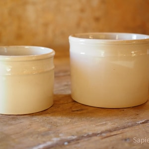 May include: Two off-white ceramic pots with a cylindrical shape. The pots have a slightly aged appearance, with a smooth, glossy finish. The smaller pot is on the left, and the larger pot is on the right. They are sitting on a wooden surface.