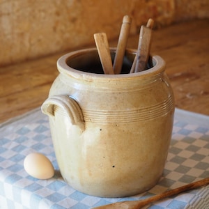 Peut inclure: Un pot en céramique beige avec une poignée, contenant des ustensiles en bois. Un œuf et une cuillère en bois reposent sur un tissu à carreaux bleu et blanc. Le pot a des rainures horizontales.