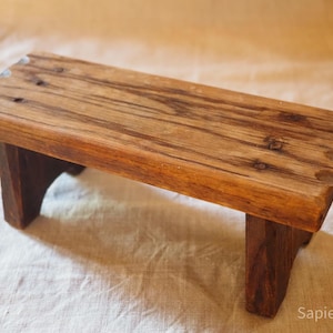Puede incluir: Un pequeño taburete de madera rectangular con un acabado natural. El taburete tiene un diseño sencillo con una parte superior plana y cuatro patas robustas. La veta de la madera es visible y el taburete parece hecho a mano. El taburete mide unos 30 cm de largo.