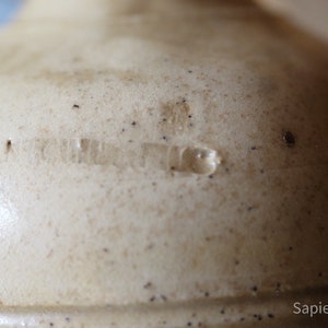 May include: Close-up of a cream-colored ceramic crock with a brown glaze and a faint inscription that reads "United States".