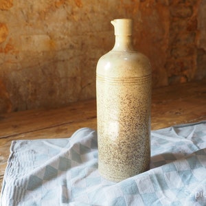 May include: A tall, cream-colored ceramic bottle with a speckled brown glaze. The bottle has a wide mouth and a rounded bottom. It is sitting on a blue and white checkered tablecloth.