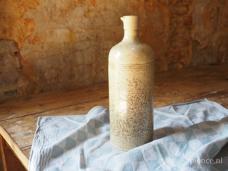 May include: A tall, cream-colored ceramic bottle with a speckled brown glaze. The bottle has a wide mouth and a rounded bottom. It is sitting on a blue and white checkered tablecloth.