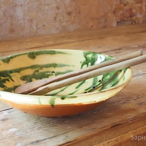 Könnte beinhalten: Eine flache Keramikschale mit cremefarbenem Inneren, grünen Farbstreifen und kleinen Abflusslöchern, kombiniert mit zwei hölzernen Servierutensilien. Die Außenseite der Schale hat eine einfarbige, terrakottabraune Oberfläche.