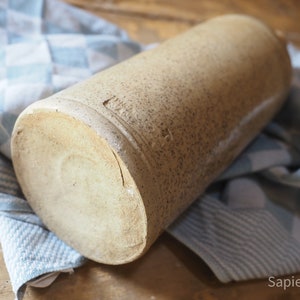 May include: A large, cylindrical, stoneware bottle with a rough, textured surface. The bottle is a light brown color and has a slightly off-white rim.