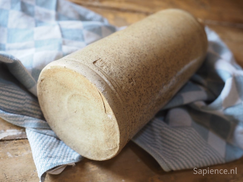 May include: A large, cylindrical, stoneware bottle with a rough, textured surface. The bottle is a light brown color and has a slightly off-white rim.