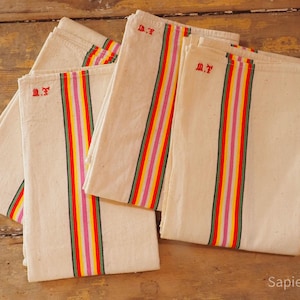 Op de afbeelding: Verschillende crèmekleurige linnen handdoeken met kleurrijke strepen in rood, geel, groen en roze. Elke handdoek heeft een rood geborduurd monogram en is netjes opgevouwen. De handdoeken liggen op een houten oppervlak.