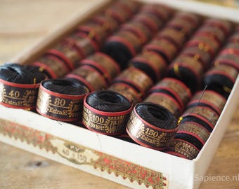 May include: A box filled with spools of black thread. The spools have decorative labels with gold lettering and red accents. The labels display numbers like 40, 80, 100, and 150, along with the text "Extra S".