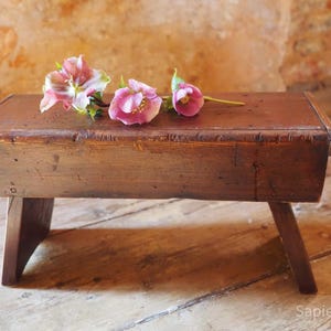 May include: A small, wooden stool with a rectangular top and three angled legs. The stool is stained a warm brown colour. Three pink and white flowers rest on the top of the stool. The background is a textured, light brown wall.