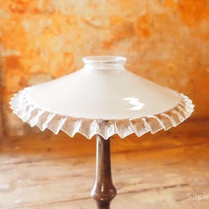 Könnte beinhalten: Vintage Tischlampe mit weißem Glasschirm und gerüschtem Rand. Die Lampe hat einen dunkelbraunen Holzfuß und steht vor einer strukturierten orangefarbenen Wand. Ein klassisches Wohnaccessoire.