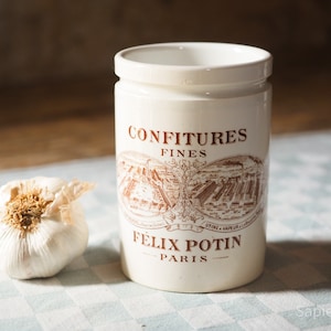 May include: White ceramic jar with a brown label that reads "Confitures Fines Felix Potin Paris". The jar is sitting on a checkered tablecloth.