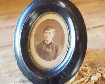 May include: An oval, black wooden picture frame with a sepia-toned portrait. The frame is placed on a wooden surface, with a strand of pearls in the foreground. The portrait features a person in a dark-colored shirt.