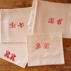 May include: Four white linen napkins with red embroidered initials. The initials are in a decorative, ornate font. The napkins are arranged on a wooden surface, with the initials "LV", "SS", "RR", and "SE" visible.