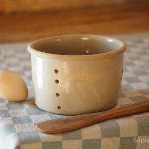 Può includere: Uno stampo per formaggio in ceramica beige con quattro piccoli fori, un uovo e un cucchiaio di legno poggiano su una tovaglia a scacchi. Lo stampo per formaggio ha una forma cilindrica con un bordo leggermente svasato. L'uovo è ovale e chiaro.