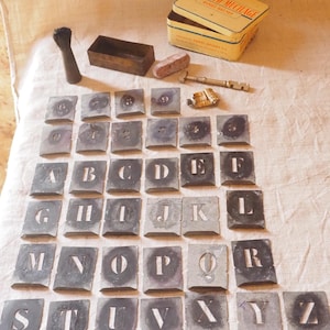 Könnte beinhalten: Eine Ansammlung von Vintage-Metallbuchstaben-Schablonen, in Reihen angeordnet, mit dem Alphabet von A bis Z. Zusätzliche Gegenstände sind ein kleiner Pinsel, eine rechteckige Box, eine Dose und Werkzeuge, alles auf einer neutralen Stoffoberfläche.