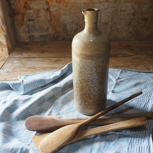 May include: A rustic, speckled ceramic bottle with a wide mouth sits on a blue and white checkered cloth with wooden spoons beside it.