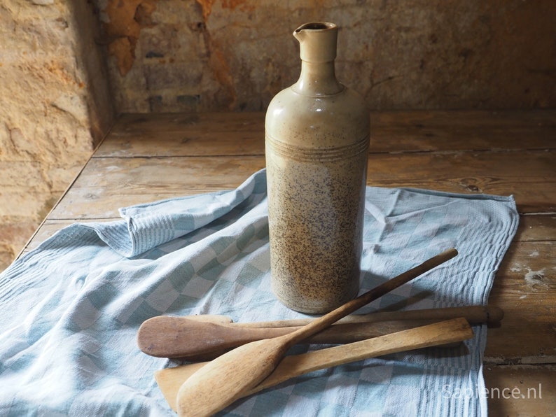 May include: A rustic, speckled ceramic bottle with a wide mouth sits on a blue and white checkered cloth with wooden spoons beside it.