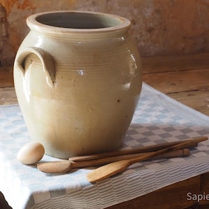 May include: A large, beige ceramic crock with two side handles, accompanied by a single light-colored egg and two wooden kitchen utensils, including a flat spatula and a long stirring stick, arranged on a light blue and white checkered cloth.