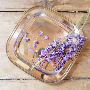Op de afbeelding: Een vierkant zilverkleurig metalen dienblad met afgeronde hoeken en een gepolijst reflecterend oppervlak. Een tros paarse blauweregenbloemen en verschillende losse bloemblaadjes zijn in het midden van het dienblad gerangschikt.
