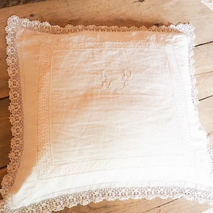 Puede incluir: Un cojín cuadrado de color crema con intrincados bordes de encaje y un diseño bordado central. El cojín presenta delicados bordes de encaje y un monograma bordado detallado, mostrando una estética vintage. El cojín está colocado sobre una superficie de madera.