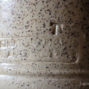 May include: Close-up of a ceramic jug with a speckled brown and white glaze. The jug has a raised inscription that reads "Erzeugniss der Wagner & Co."