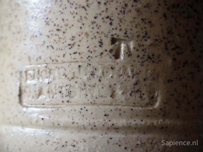 May include: Close-up of a ceramic jug with a speckled brown and white glaze. The jug has a raised inscription that reads "Erzeugniss der Wagner & Co."