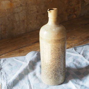 May include: A ceramic bottle with a speckled beige and brown glaze. The bottle has a narrow neck and a rounded body. It is sitting on a blue and white checkered tablecloth.