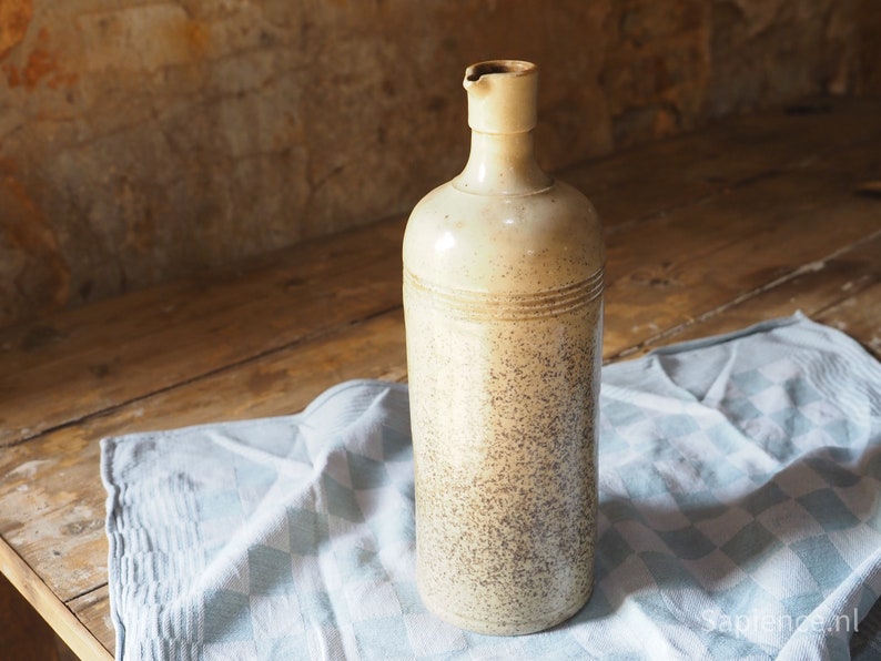 May include: A ceramic bottle with a speckled beige and brown glaze. The bottle has a narrow neck and a rounded body. It is sitting on a blue and white checkered tablecloth.
