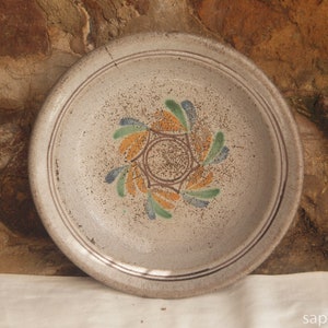 May include: A white ceramic plate with a decorative design of stylised flowers in shades of green, yellow, and orange. The plate has a raised rim and a slightly textured surface.