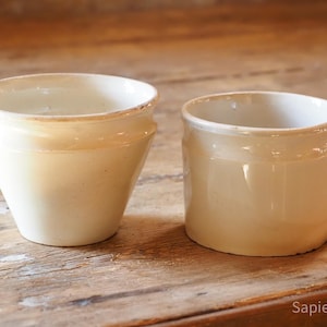 Könnte beinhalten: Zwei cremefarbene Keramiktöpfe. Einer hat eine konische Form mit leicht ausgestelltem Rand, der andere ist ein kürzerer, zylindrischer Topf. Beide haben eine glänzende Oberfläche und stehen auf einer verwitterten Holzoberfläche.