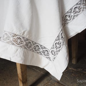 May include: Close-up of a white tablecloth with a decorative black lace border. The tablecloth is draped over a wooden table, showcasing the intricate lace design and the fabric's texture. The overall theme is home decor.
