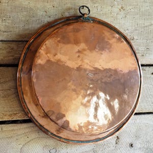May include: A round, hammered copper tray with a polished, reflective surface and a dark patina along the edges. The tray features a small, circular metal hanging loop attached to the top rim, allowing it to be displayed vertically on a wall.