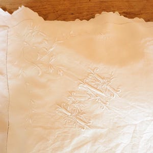 May include: Cream-colored, embroidered linen textile with a scalloped edge. The embroidery features floral and scroll designs. The fabric is slightly wrinkled, suggesting it is vintage or handmade. The overall aesthetic is delicate and elegant.