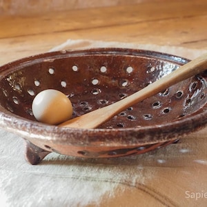 May include: A round, brown, glazed ceramic colander with small drainage holes, three short feet, and a wide rim. Inside the bowl sits a single smooth, off-white egg and a long, light-coloured wooden spoon resting diagonally across the basin.