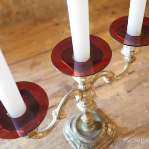 May include: A gold-colored candelabra with three arms, each holding a white candle. The candle holders have a red glass base. The candelabra is on a wooden surface.
