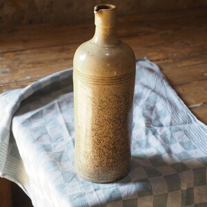 May include: A ceramic bottle with a speckled brown and beige glaze. The bottle has a wide mouth and a rounded body. It is sitting on a white and blue checkered tablecloth.