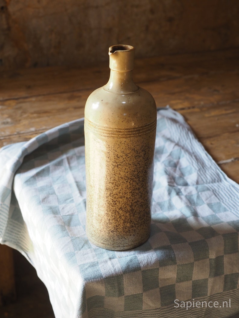 May include: A ceramic bottle with a speckled brown and beige glaze. The bottle has a wide mouth and a rounded body. It is sitting on a white and blue checkered tablecloth.