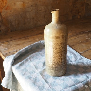 May include: A ceramic bottle with a speckled brown and beige glaze. The bottle has a wide mouth and a rounded body. It is sitting on a blue and white checkered tablecloth.