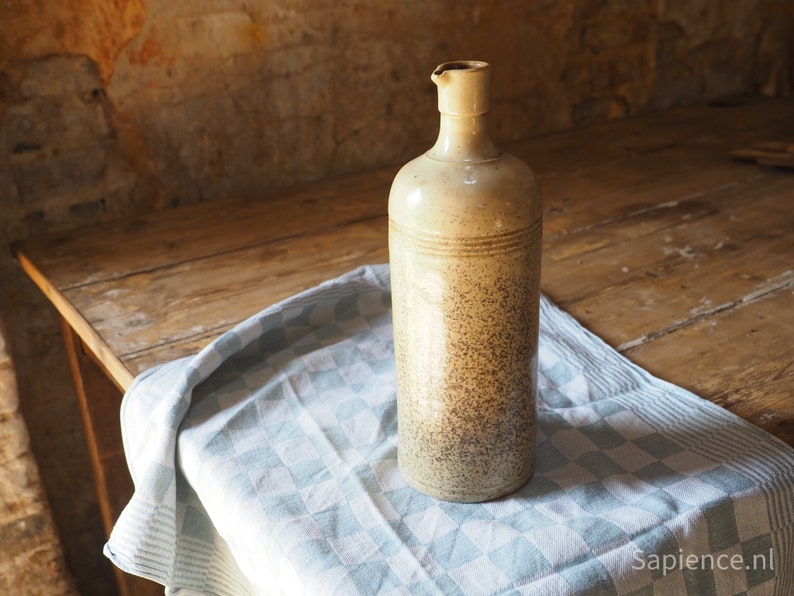 May include: A ceramic bottle with a speckled brown and beige glaze. The bottle has a wide mouth and a rounded body. It is sitting on a blue and white checkered tablecloth.