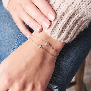 May include: A delicate silver chain bracelet with a small diamond heart and star charm. The bracelet is worn on a person's wrist.