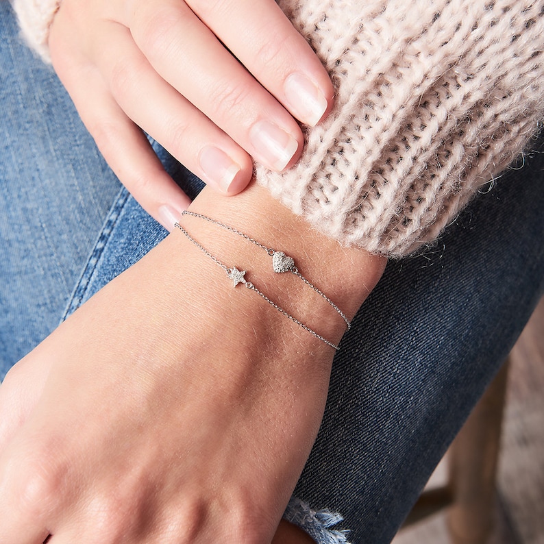 May include: A delicate silver chain bracelet with a small diamond heart and star charm. The bracelet is worn on a person's wrist.