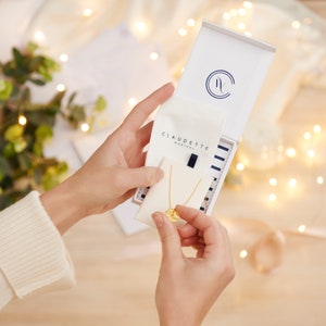May include: A white box with a blue and white striped interior, containing a gold necklace with a round pendant. The box has the text "CLAUDETTE MONTREAL" printed on it.