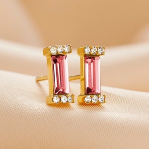 May include: A pair of gold-colored stud earrings. Each earring features a rectangular, pink gemstone center, framed by small, clear crystals. The earrings are set against a soft, peach-colored background.