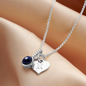 Pode incluir: Um colar de prata com corrente e três pingentes. Inclui uma pedra preciosa azul escura, um pingente em forma de coração com a letra "H" gravada e uma pequena peça decorativa com pedras claras.