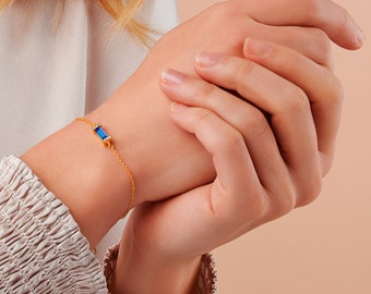 May include: A gold chain bracelet with a blue rectangular gemstone set in a gold frame. The gemstone is surrounded by smaller clear gemstones.