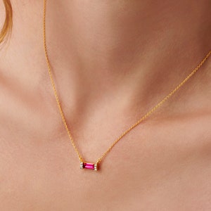 以下が含まれることがあります： 小さなクリアストーンでアクセントが付けられた、小さなピンクの四角形の宝石のペンダントが付いた、繊細なゴールドチェーンネックレスです。