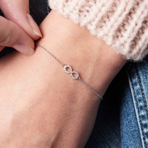 Könnte beinhalten: Ein silbernes Armband mit einem Unendlichkeitszeichen und kleinen, klaren Steinen. Das Armband befindet sich an einem Handgelenk.